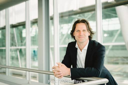 Auf ein Geländer aufgestützt lächelt Timo Leukefeld im Jacket mit weißem Hemd und offenem Kragen in die Kamera. Im Hintergrund Glas.