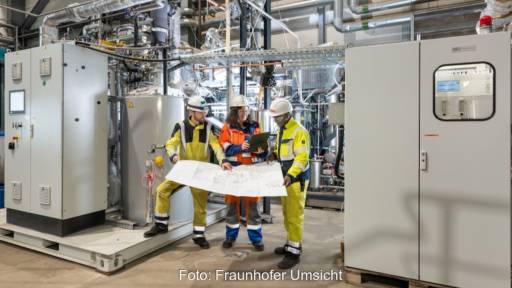 Blick in die Pyrolyse-Anlage bei RWE: Zwei Männer mit gelben Schutzanzügen und eine Frau mit orangefarbenem Schutzanzug stehen zwischen grauen Gehäusen und halten einen Plan in der Hand.