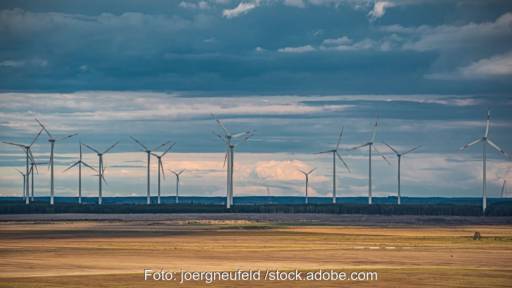 Eine Reihe großer Windenergie-Anlagen vor Gewitterhimmel, im Vordergrund ein Stoppelfeld. Symbolbild für Windenergie, Windpark.