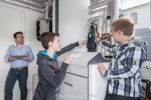 Zwei Jungs freuen sich im Heizungskeller über eine Wärmepumpe; der Vater steht mit einem Laptop lachend daneben.