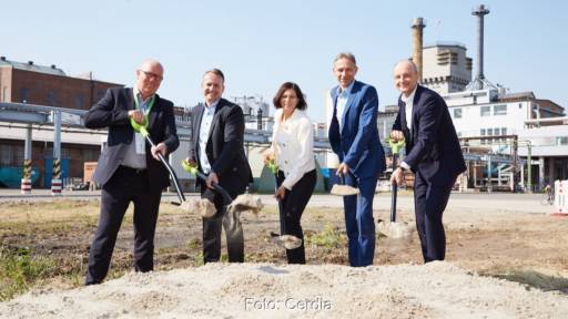 Vier Männer und eine Frau mit Schaufeln, im Hintergrund eine Fabrik: Spatenstich für Biomasse-Heizwerk.