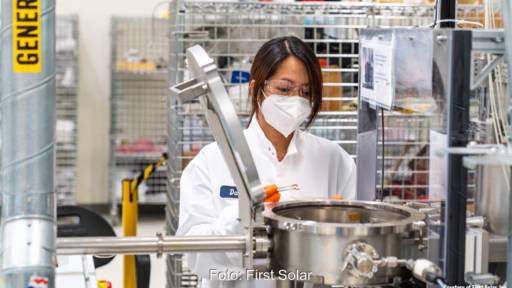 Eine Frau mit weißem Kittel und Mundschutz vor einer Labor-Apparatur hält eine kleine Solarzelle in einer Pinzette - Forschung an Dünnschicht Photovoltaik bei First Solar