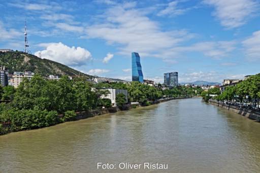 Ein breiter Fluss durchfließt die Stadt Tiflis: an den Ufern grüne Vegetation, Häuser und Hochhäuser im Hintergrund.