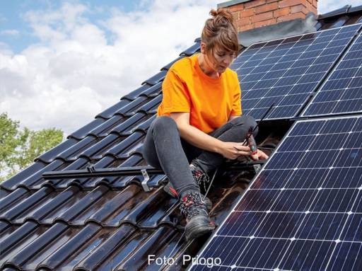 Installateurin montiert sitzend auf einem Schrägdach Solarmodule.