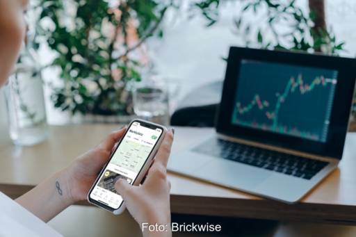 Zwei Hände halten ein Smartphone, Computerbildschirm mit Kursverlauf: Blockchain ermöglicht Bürgerbeteiligung per App