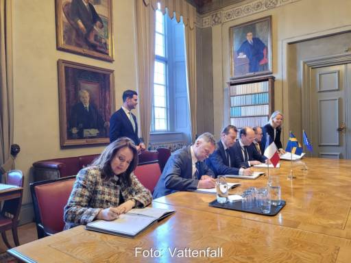 Menschen sitzen an breitem Holztisch auf Lederstühlen und unterschreiben Dokumente.