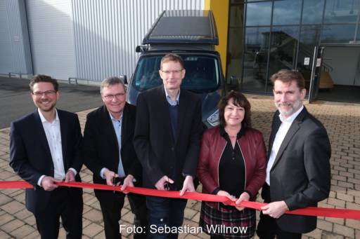 Menschen in Anzügen schneiden rotes Band durch, im Hintergrund Fahrzeug mit PV-Dach.