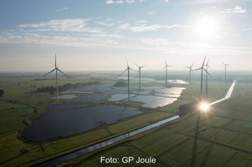 Luftbild zeigt großen PV-Park unter Windenergieanlagen im grünen Flachland.