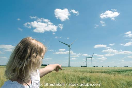 Eine junge Frau zeigt Windkraftanlagen auf einer grünen Wiese vor blauem Himmel als Zeichen für Bürgerenergie.
