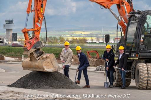 Mehrere Männer im Anzug und Schutzhelm bei einem symbolischen Spatenstich neben einem Bagger.