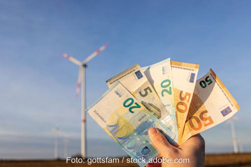 Hand hält Geldscheine vor Windpark in die Höche - Symbol für finanzielle Beteiligung von Kommunen
