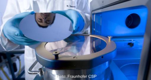 Eine Hand legt eine glänzende Scheibe auf einen Glasteller, ein Gesicht spiegelt sich in der Scheibe - Laborszene am Fraunhofer CSP.