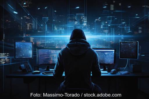 In einem dunklen Raum sitzt ein Mensch mit Kaputzenpulli vor Monitoren.