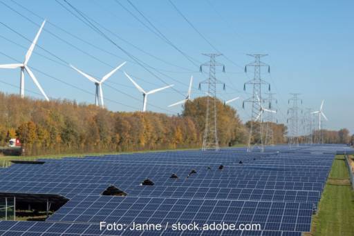Im Vordergrund eine dunkel schimmernde Freiflächen-Photovoltaikanlage, dahinter Bäume, hinter denen wiederum die weißen Flügel von Windkraftanlagen sichtbar sind. In der Mitte und oben rechts im Bild sind Strommasten zu sehen. Ein Symbolbild für den Artikel, in dem es um neue Regeln für Stromnetzentgelte geht.