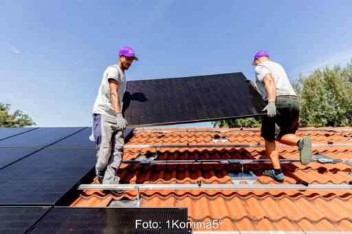 Zwei Monteure tragen ein PV-Modul auf ein mit roten Dachschindeln gedecktes Dach.