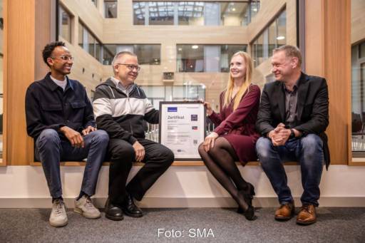 Vier Personen in einem Bürogebäude sitzend mit Zertifikatsurkunde.