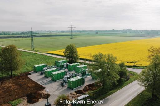 An einer Straße vor Feldern steht eine Anlage mit grünen Containern. Sie enthalten große Stromspeicher