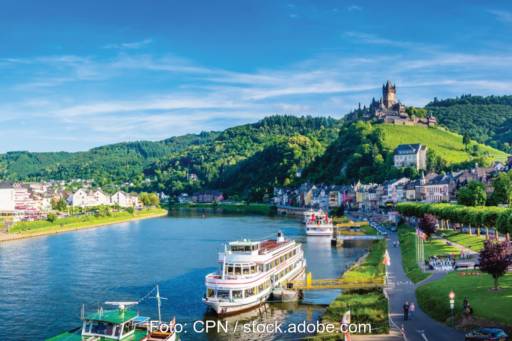 Die Mosel mit Hügeln und einer Burg im Hintergrund. Auf den Fluss sind Schiffe zu sehen, die an einer Anlegestelle liegen.