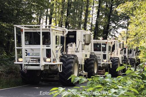 Weiß lackierte Kraftwagen mit großen Reifen in einer Reihe auf einem Waldweg. Vibro-Trucks erkunden den Untergrund, um das Potenzial für Tiefengeothermie-Bohrungen zu erkunden.