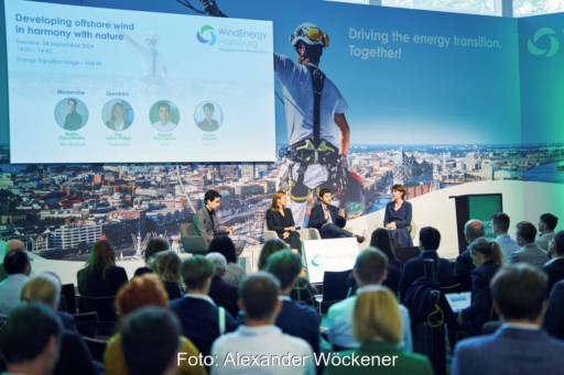 Konferenz mit vier Personen auf dem Podium und Fotos der Windenergie.