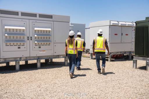 Drei Personen in Sicherheitswesten gehen durch einen Batteriepark unter blauem Himmel.