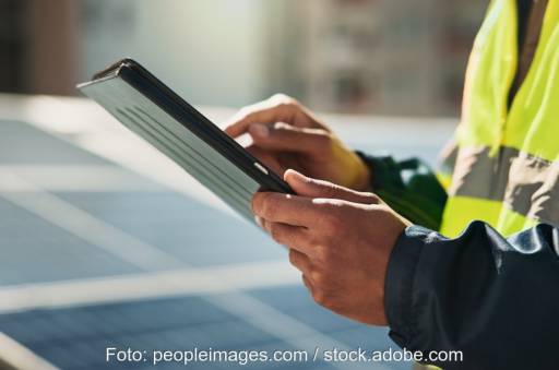 Zwei Hände halten ein Tablett und arbeiten daran - im Hintergrund eine Photovoltaikanlage. Symbolbild zur Digitalisierung des Betriebs von PV-Anlagen.