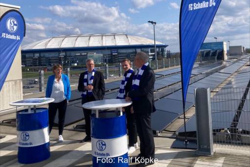 Vier Personen stehen zwischen Bannern des Fußballereins Schalke 04 vor einer Photovoltaikanlage in Gelsenkirchen. Im Hintergrund ist das Fußballstadion.