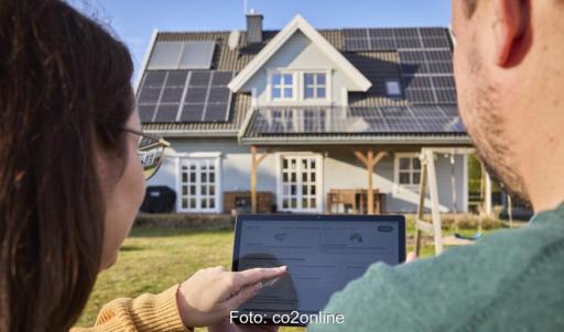 Zwei Menschen schauen vor Haus mit Photovoltaik-Anlage auf ein Tablett, diese lohnt sich auch 2025.