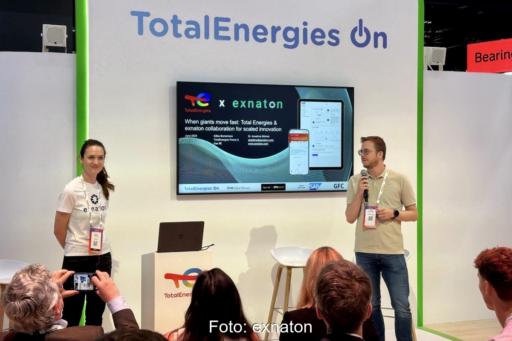 Auf einem Messe-Stand von TotalEnerges stehen die beiden Verantwortlichen vor einem Monitor und Publikum.