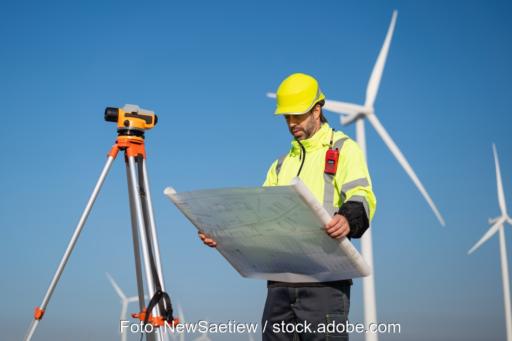 Das Foto zeigt einen Menschen in gelber Jacke mit gelber Kappe, der einen Plan in der Hand hält. Neben ihm ein Vermessungsgerät, im Hintergrund Windkraftanlagen (Beschleunigungsgebiete RED III)