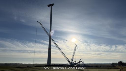 Vor einer milchigen Sonne finden Arbeiten mit einem Kran an einer Windenergieanlage statt.