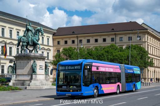 Ein knallblauer Elektro-Bus mit pinkem Element fährt vor historischen Gebäuden in München entlang.