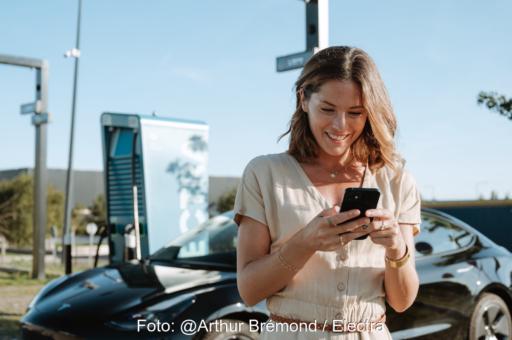 Eine Frau schaut an einer Ladesäule auf ihr Handy.