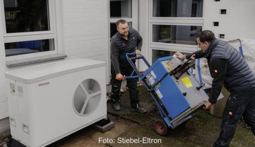 Heizungstausch: Zwei Monteure ziehen ein blaues Gerät auf einer Sackkarre aus einem Haus, draußen steht eine weiße Luft-Wärmepumpe.