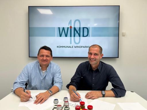 Zwei Personen sitzen hinter einem weißen Tisch und vor einer Bildschirm-Projektion an der Wand mit Papieren vor sich.