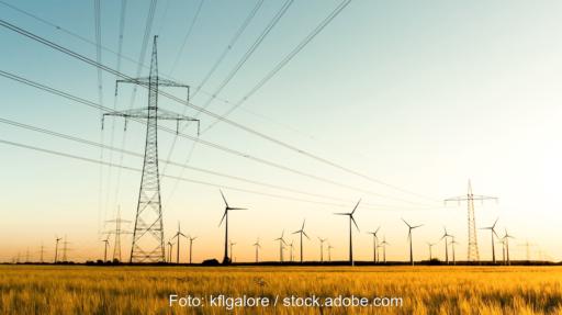 Strommasten auf einem Feld, im Hintergrund Windkraftanlagen - Versorgungssicherheit auch mit erneuerbaren Energien.