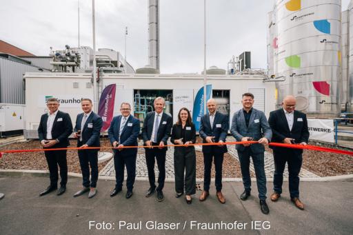 Mehrere Personen stehen draußen vor der Anlage mit einer Schere vor einem roten Band.