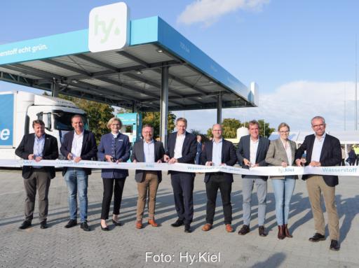 Mehrere Personen stehen draußen vor der Wasserstofftankstelle mit einer Schere vor einem hellen Band.