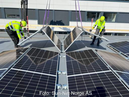 Zwei Personen auf einem Dach falten eine Einheit aus vier Photovoltaik-Modulen auseinander, die an einem Kran hängen.