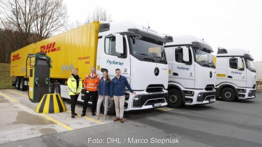 Vier Personen stehen neben einer weißen eActros-Zugmaschine plus einem gelbem DHL-Anhänger. Schräg dahinter zwei weitere E-LKW.