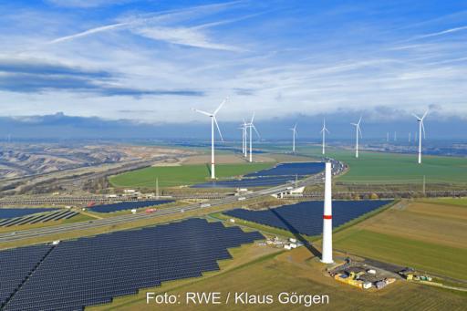 Ein Luftbild von schräg vorn mit Horizont: links oben Mitte der Tagebau Garzweiler, von links unten kommend die A44n mit Photovoltaikanlagen zu beiden Seiten sowie Windenergieanlagen dazwischen.