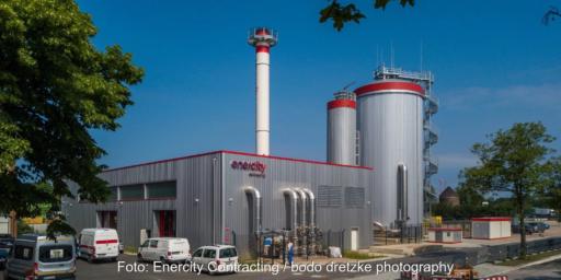 Gebäude einer Energiezentrale unter blauem Himmel und historischem Wasserspeicher im Hintergrund.