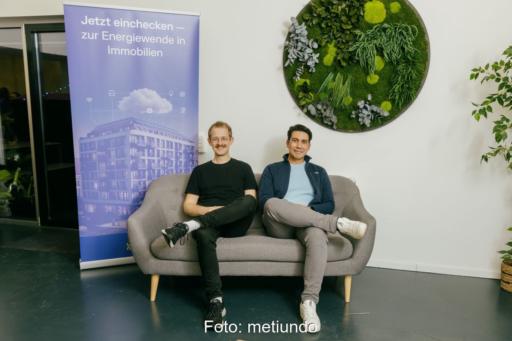 Zwei Männer in legerer Kleidung auf einem Sofa im Büroumfeld.