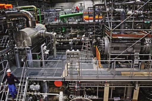 Blick in eine große Wärmepumpe mit vielen Leitungen und weiteren Komponenten. Gitterroste erschließen Arbeitsebenen. Darauf stehen Menschen mit Sicherheitshelm.