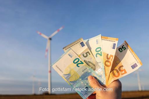 Eine Hand hält Geldscheine vor Windenergieanlage, die Redispatch-Abrechnung erfolgt ab 2026 direkt zwischen Anlagenbetreiber und Netzbetreiber.