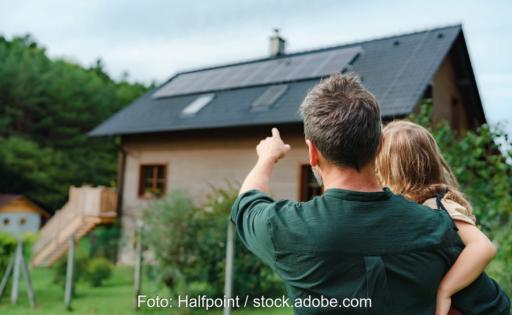 Ein Mann mit Kind im Arm vor Haus mit Photovoltaik-Anlage.