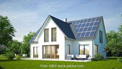 Weißes Einfamilienhaus, Neubau, mit Photovoltaik-Anlage und grünem Rasen vor blauem Himmel.