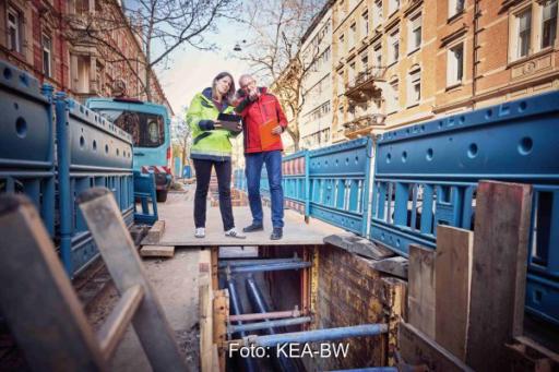 Das Foto zeigt zwei Personen stehend in einer Straßenschlucht mit mehrstöckigen Altbauten rechts und links. Unter ihren Füßen eine Baustelle, die Straße ist aufgerissen und legt Rohre frei.