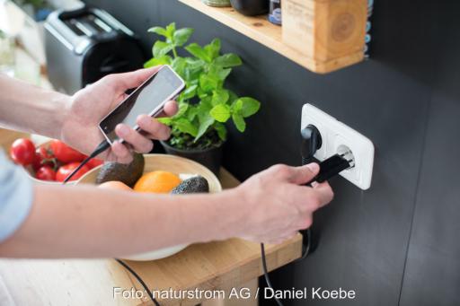 Zu sehen ist in einem Küchenumfeld mit Toaster, Basilikum und Obstkob wie eine linke Hand ein Smartphone hält und eine rechte Hand das Ladekabel in eine weiße Steckdose steckt.