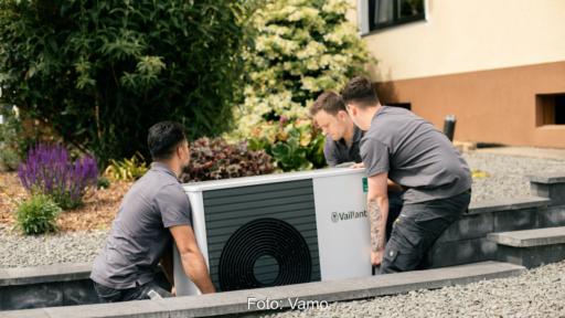 Das Bild zeigt drei grau gekleidete Männer in Arbeitskleidung, die gemeinsam zu einem Haus eine Wärmepumpe tragen.